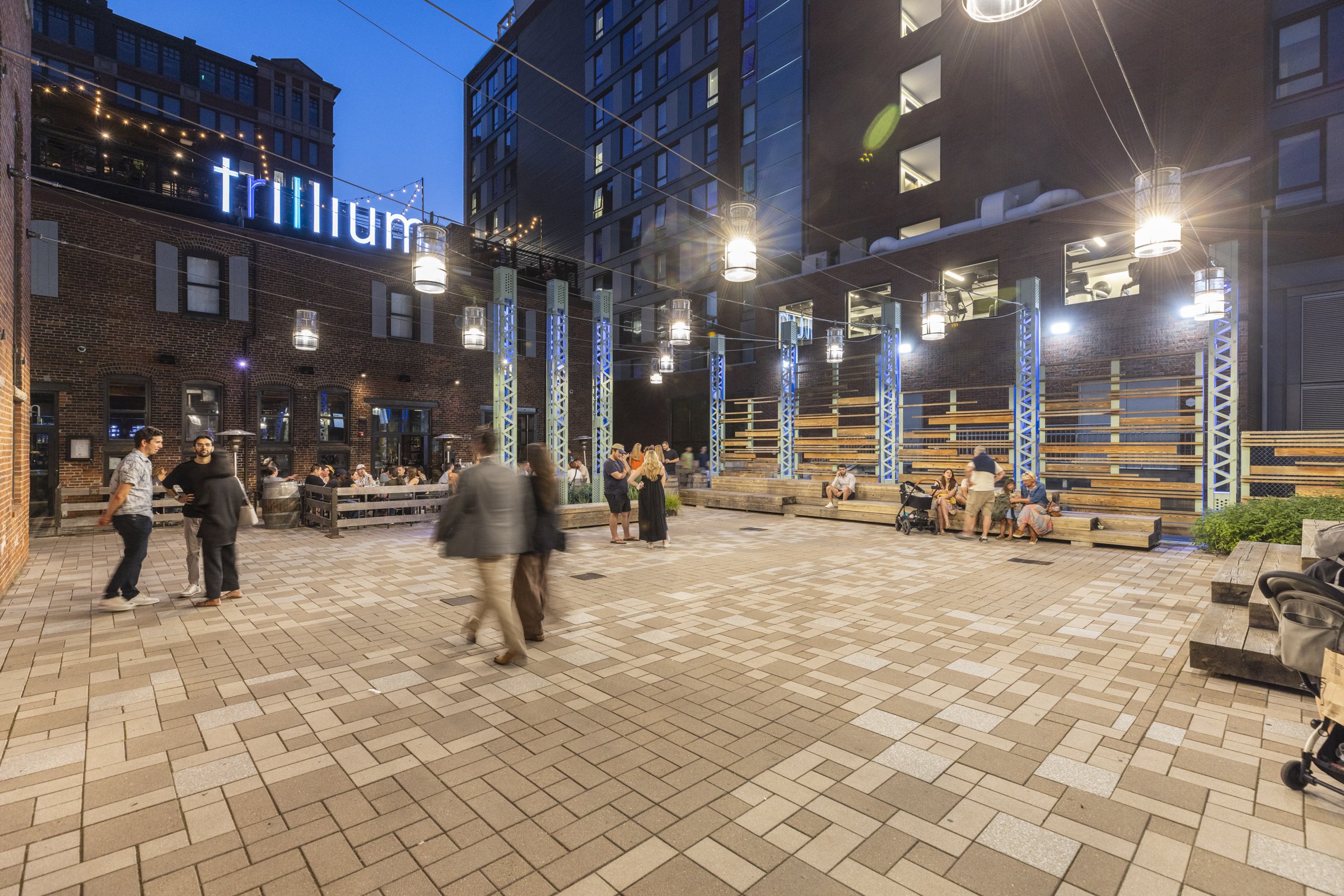 Outdoor urban patio at dusk with people socializing, wooden benches, string lights, modern building with a sign reading 'trillium' illuminated in blue.