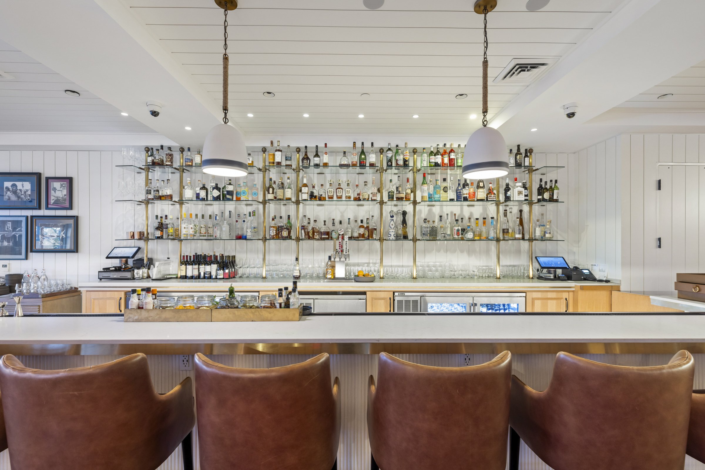 Interior of a bar with bottles of liquor displayed on metal shelves behind the counter. There are two hanging pendant lights and four brown leather chairs at the bar.