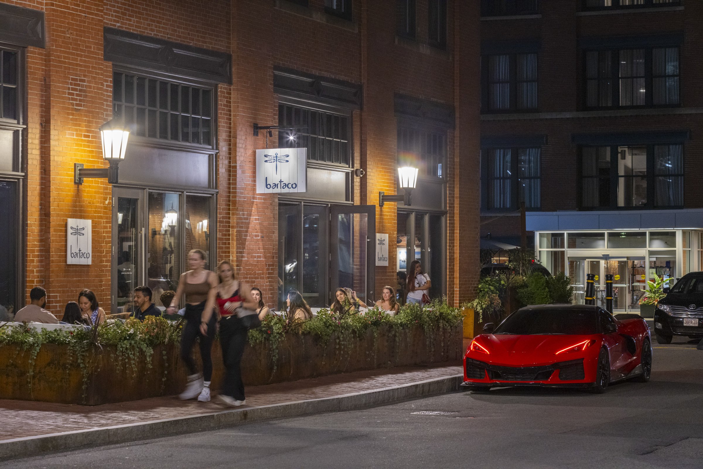 Nighttime city street scene with a lit brick restaurant called 'Bar Tacos' with people dining outside, two women walking past, and a red sports car parked on the street.