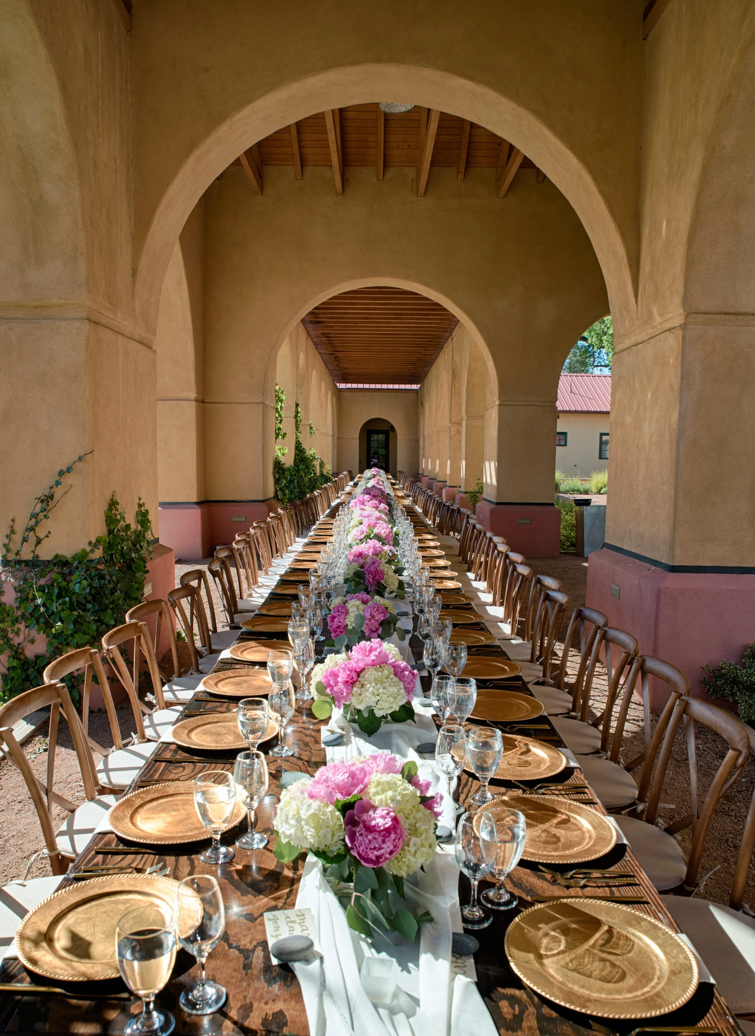 Long banquet table set with pink and white flower centerpieces, gold chargers, water glasses, and cutlery under archways with a wooden ceiling, with outdoor scenery visible.