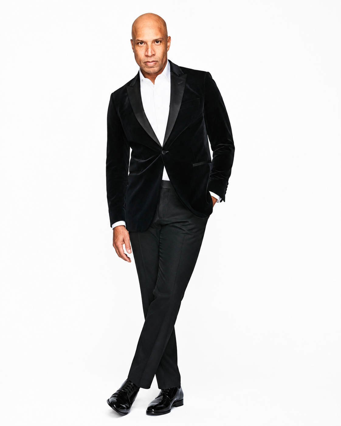 A man in a black velvet tuxedo with a white shirt, black dress pants, and shiny black dress shoes standing against a plain white background.