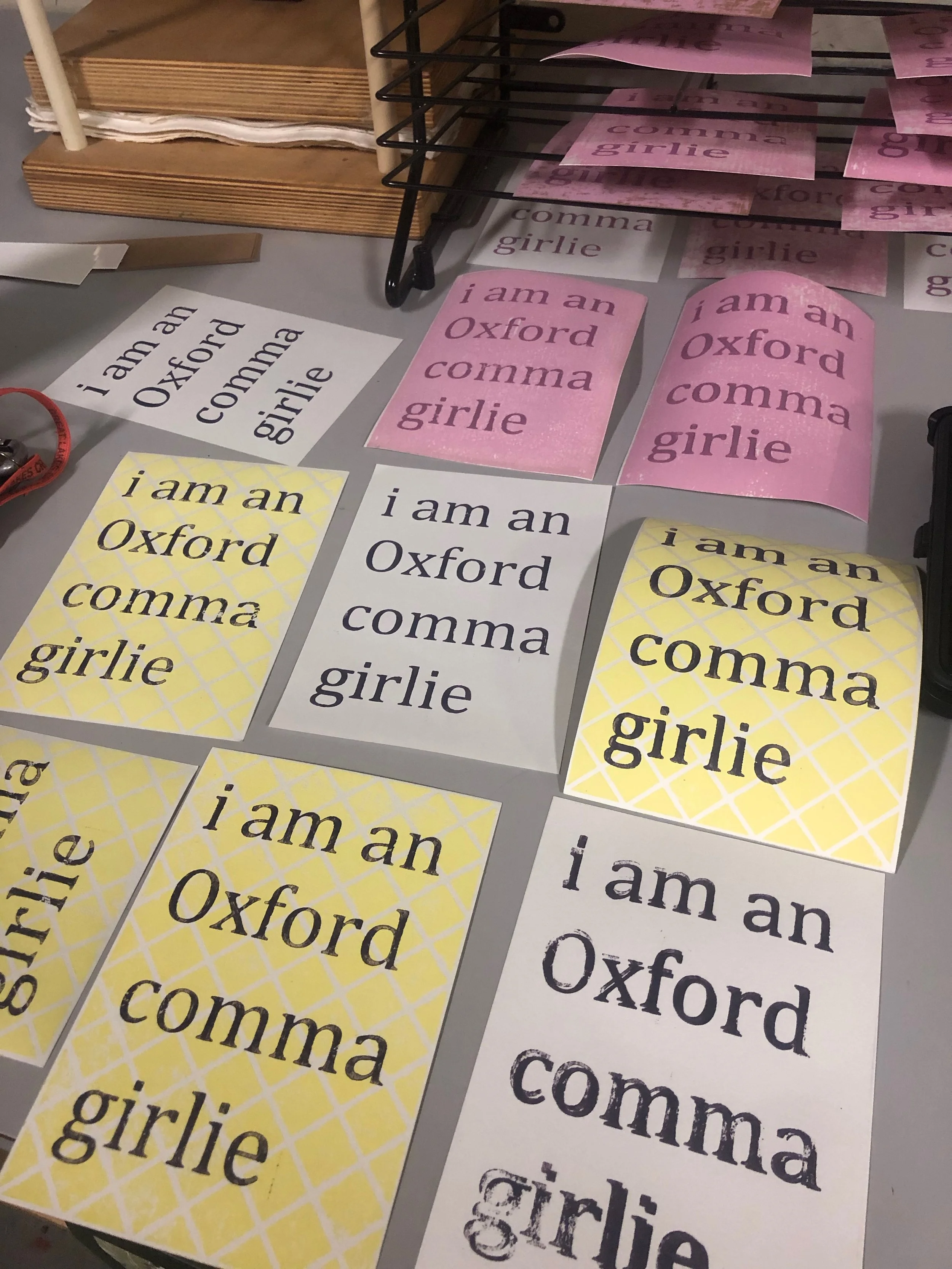 Several posters are spread out on a table in different color cobinations read "I am an Oxford comma girlie"