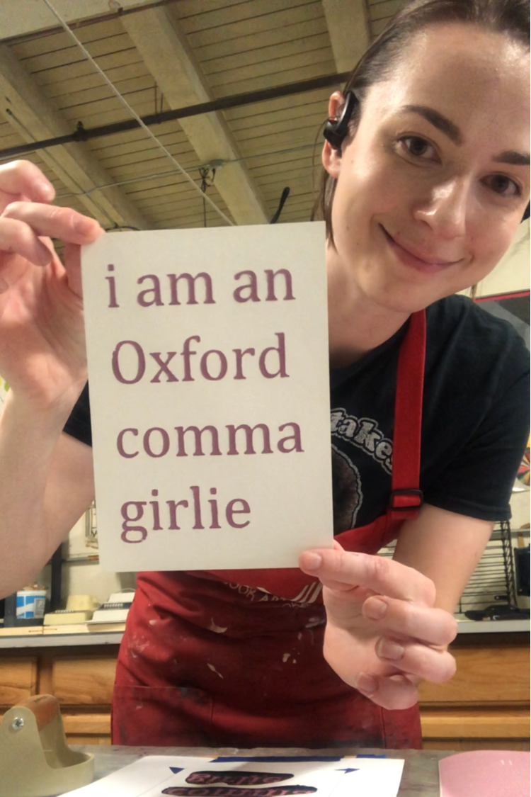 A woman holds up a finished poster that reads "I am an Oxford comma girlie"