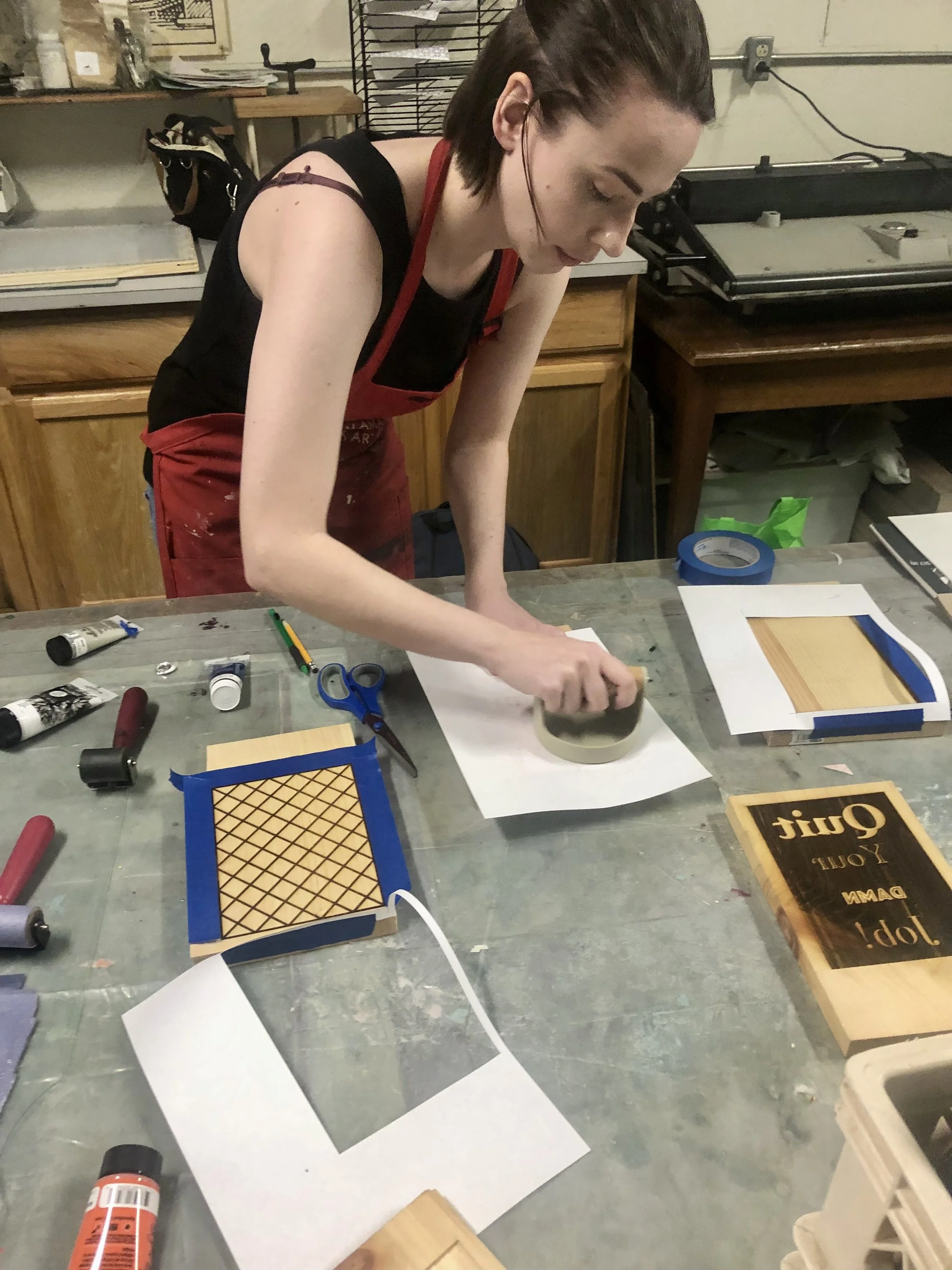 A woman stands at a table putting pressure on the printing plate to make a print.