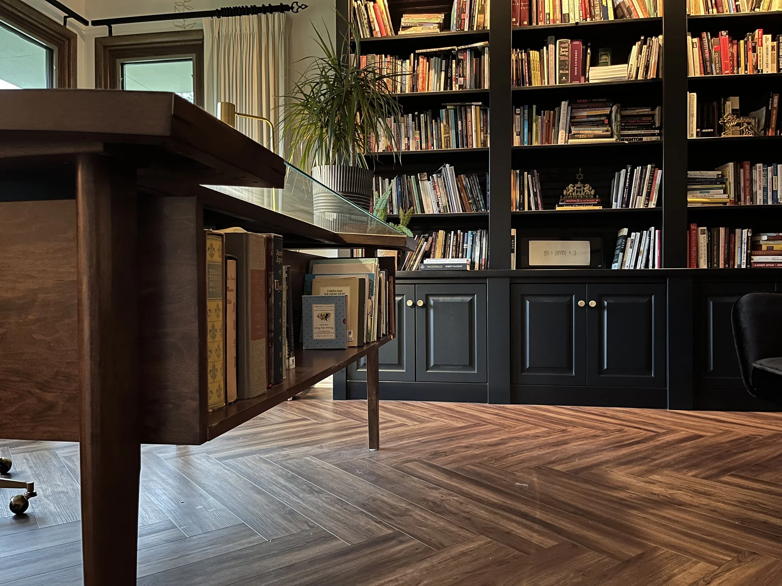 Interior view of a cozy home library with black bookshelves filled with colorful books, a wooden table, several potted plants, and large windows with curtains letting in natural light.