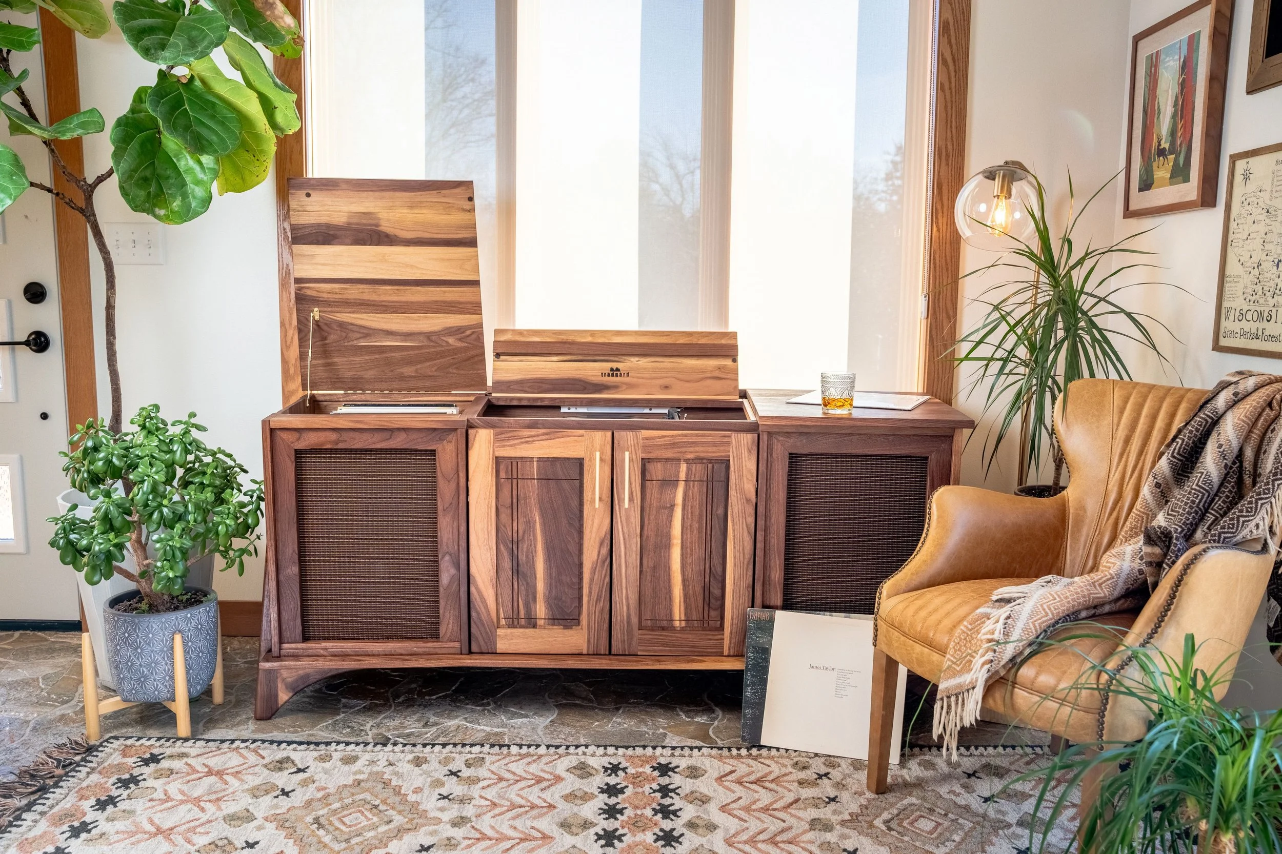 Living room corner with a wooden sideboard, large plants, artwork on the walls, a vintage record player, a glass of whiskey on the sideboard, and a mustard-colored armchair with a blanket, on a patterned rug.