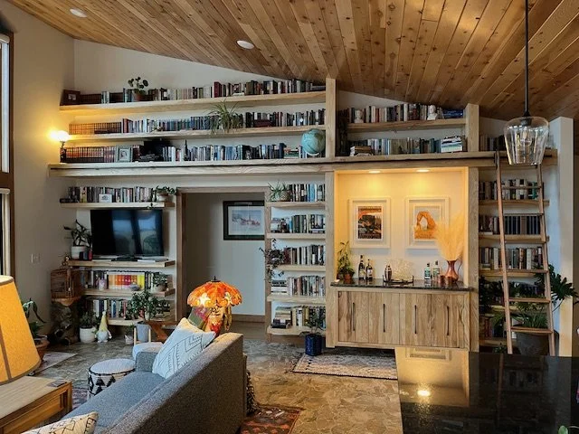 Living room with built-in bookshelves filled with books and decor, an open doorway, a gray sofa with decorative pillows, a colorful lamp, and framed artwork on the wall, with a sloped wooden ceiling.