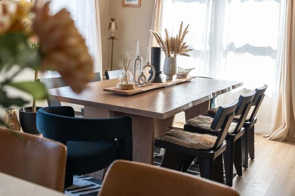Wood dining table with decorative vases, dried floral arrangement, and small ornaments, surrounded by black and tan chairs in a well-lit room.