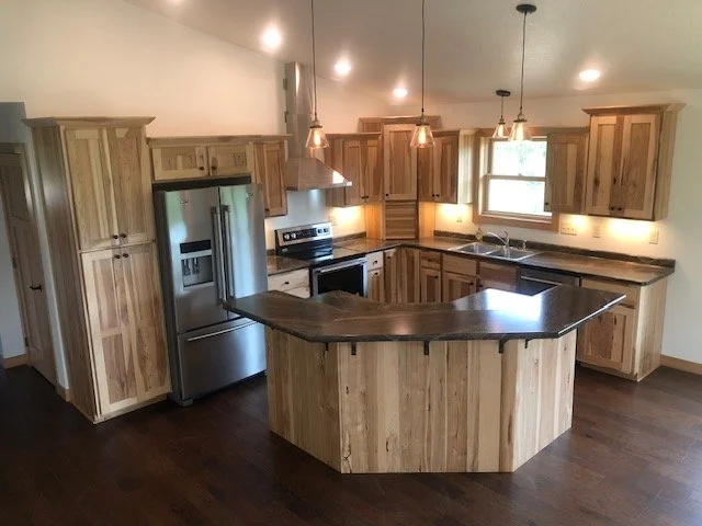 Kitchen with wooden cabinets, stainless steel refrigerator, oven, and black countertops, featuring a central island with seating area and pendant lighting.