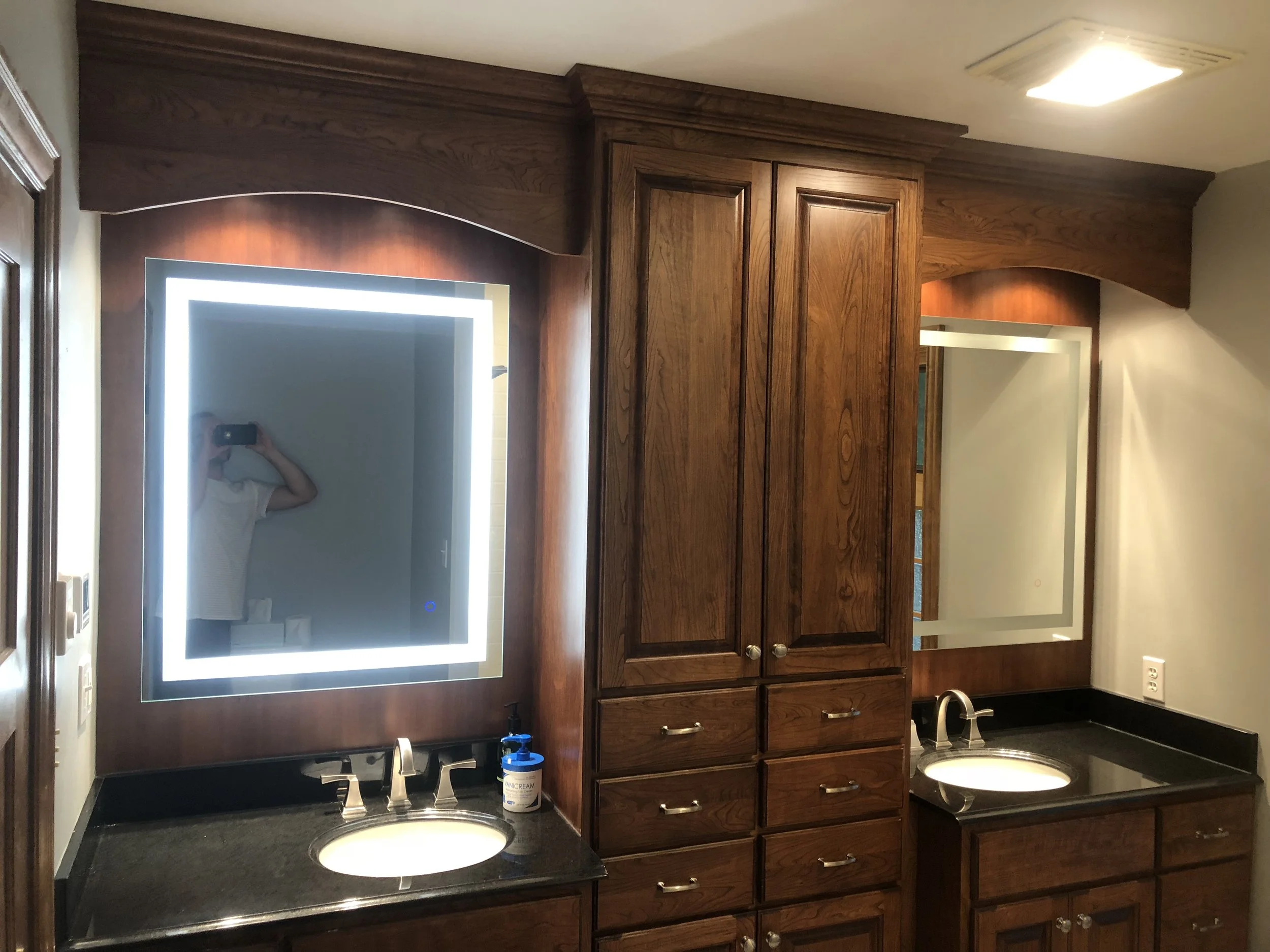 Bathroom vanity with a large illuminated mirror, wooden cabinets, a sink, and bottles of hand soap on the countertop.
