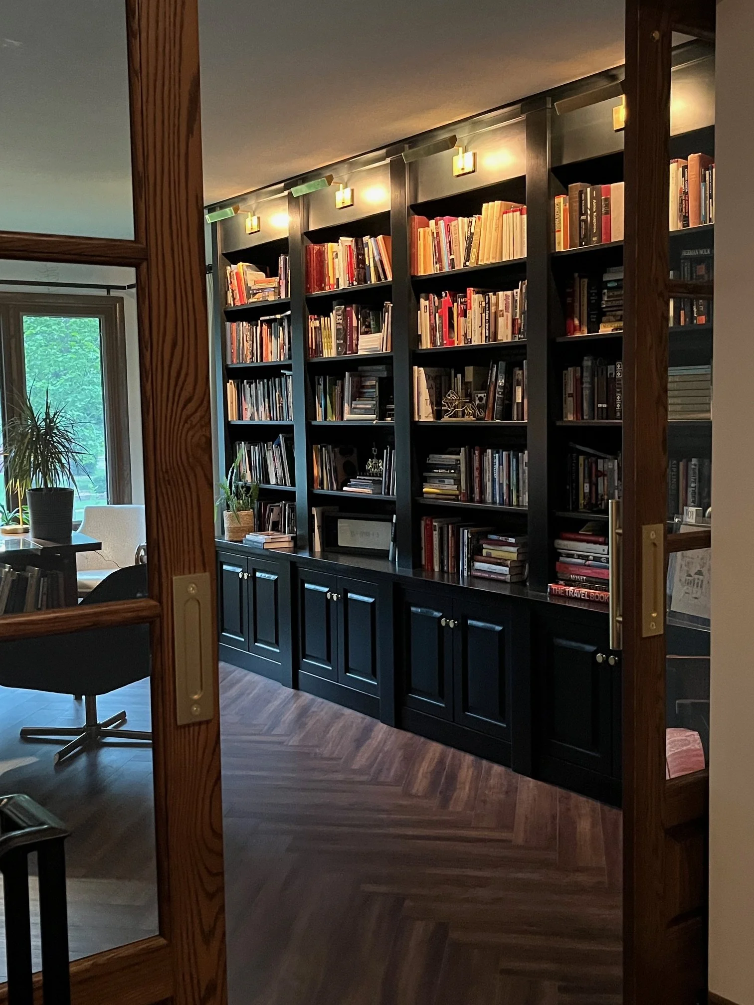 A black bookshelf filled with books inside a room, viewed through a wooden door frame, with a window and plants in the background.