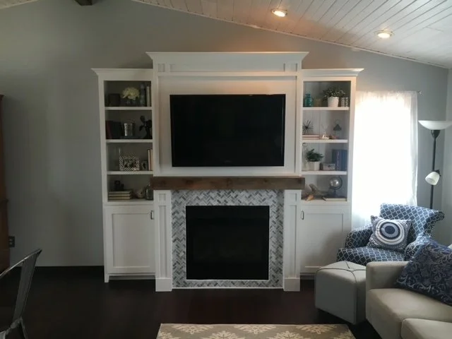 Living room with a built-in white entertainment center, a flat-screen TV, a fireplace with tiled surround, and a gray armchair with decorative pillows next to a tall floor lamp.