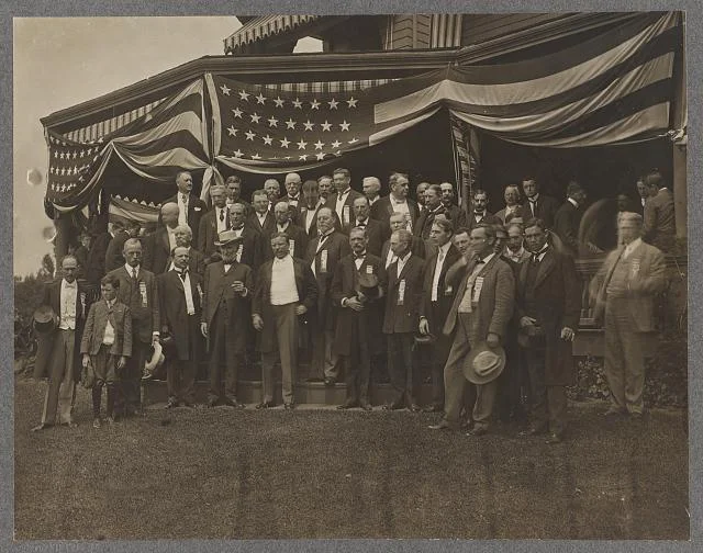 Teddy Roosevelt Standing in Center of a Group of Men Outside a Porch Draped with Flags | Circa 1904