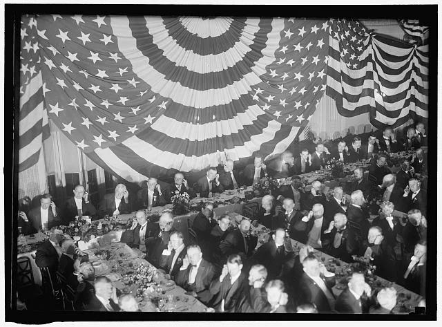 Banquet Scene with Flag Draping and Men Seated Holding Up Listening Device to One Ear | Circa 1913