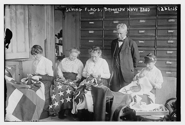Women Hand Sewing an American Flag and Other Flags as a Man Watches at the Brooklyn Navy Yard | Circa 1917