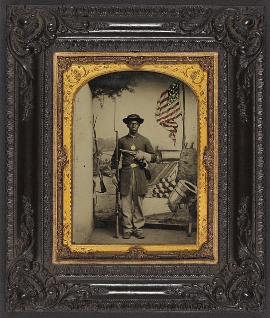 African American Soldier in Union Uniform with a Rifle and Revolver in Front Painted Backdrop Showing Weapons and American Flag at Benton Barracks, Saint Louis, Missouri | Circa 1863-1865