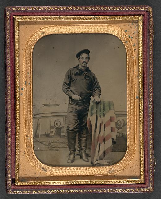 Sailor in Union Uniform Resting Hands on American Flag-Draped Table in Front of Painted Backdrop Showing Naval Scene | Circa 1861-1865