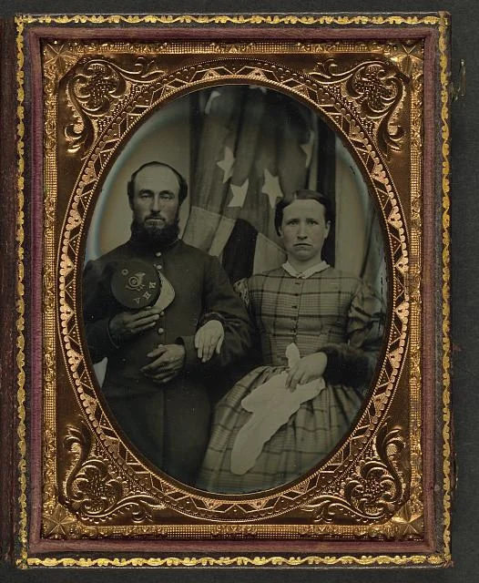 Soldier in Union Frock Coat Holding Company G, 12th New Hampshire Infantry Regiment Forage Cap Next to Women in Front of the American Flag | Circa 1861-1865