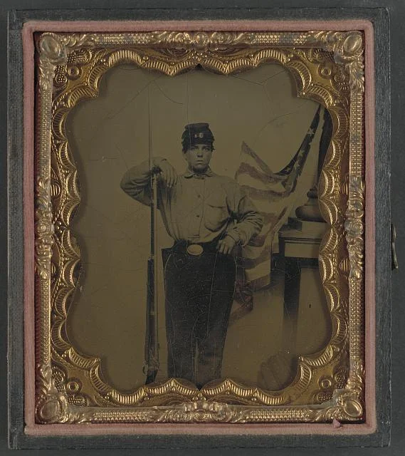 Soldier in Union Uniform with Bayoneted Musket in Front of Painted Backdrop Showing American Flag and Column Pedestal | Circa 1861-1865