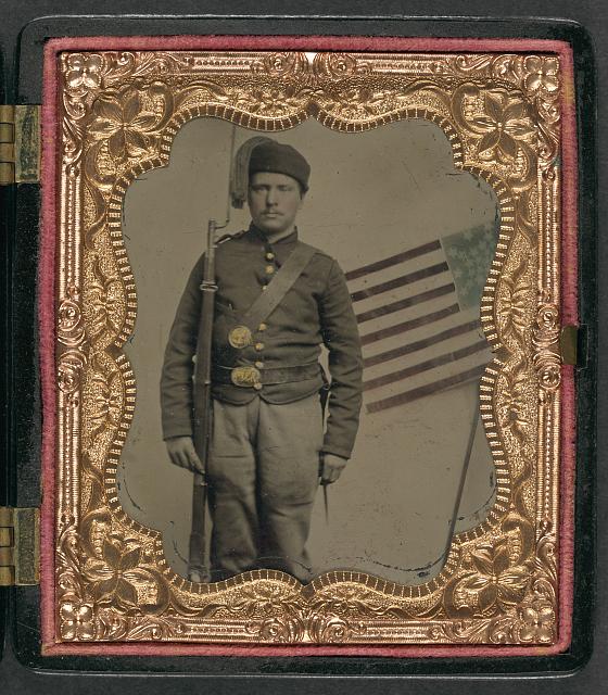Soldier in Union Uniform with Bayoneted Musket in Front of Painted Backdrop Showing American Flag | Circa 1861-1865