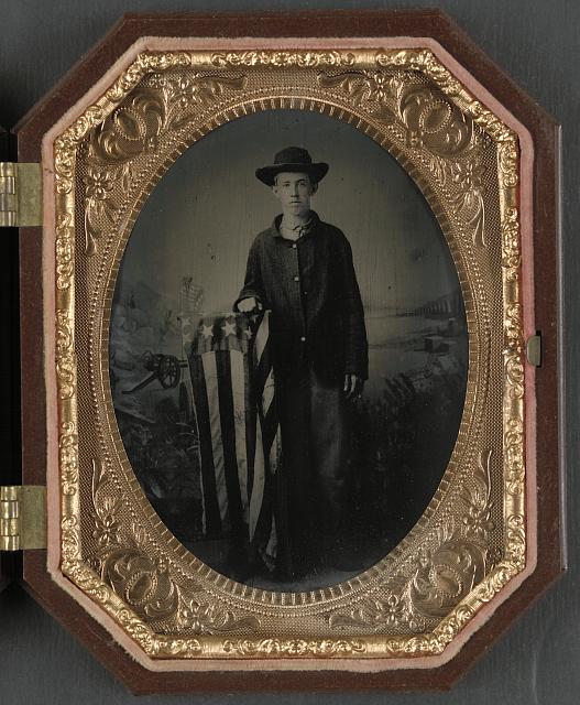 Young Soldier in Union Uniform with American Flag-Draped Stand in Front of Backdrop Showing Battlefield Scene | Circa 1861-1865