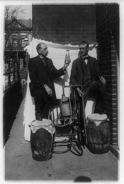 Two Men Standing Outdoors with Small Still, One of Them Holding Up a Bottle of Liquor | Circa 1921