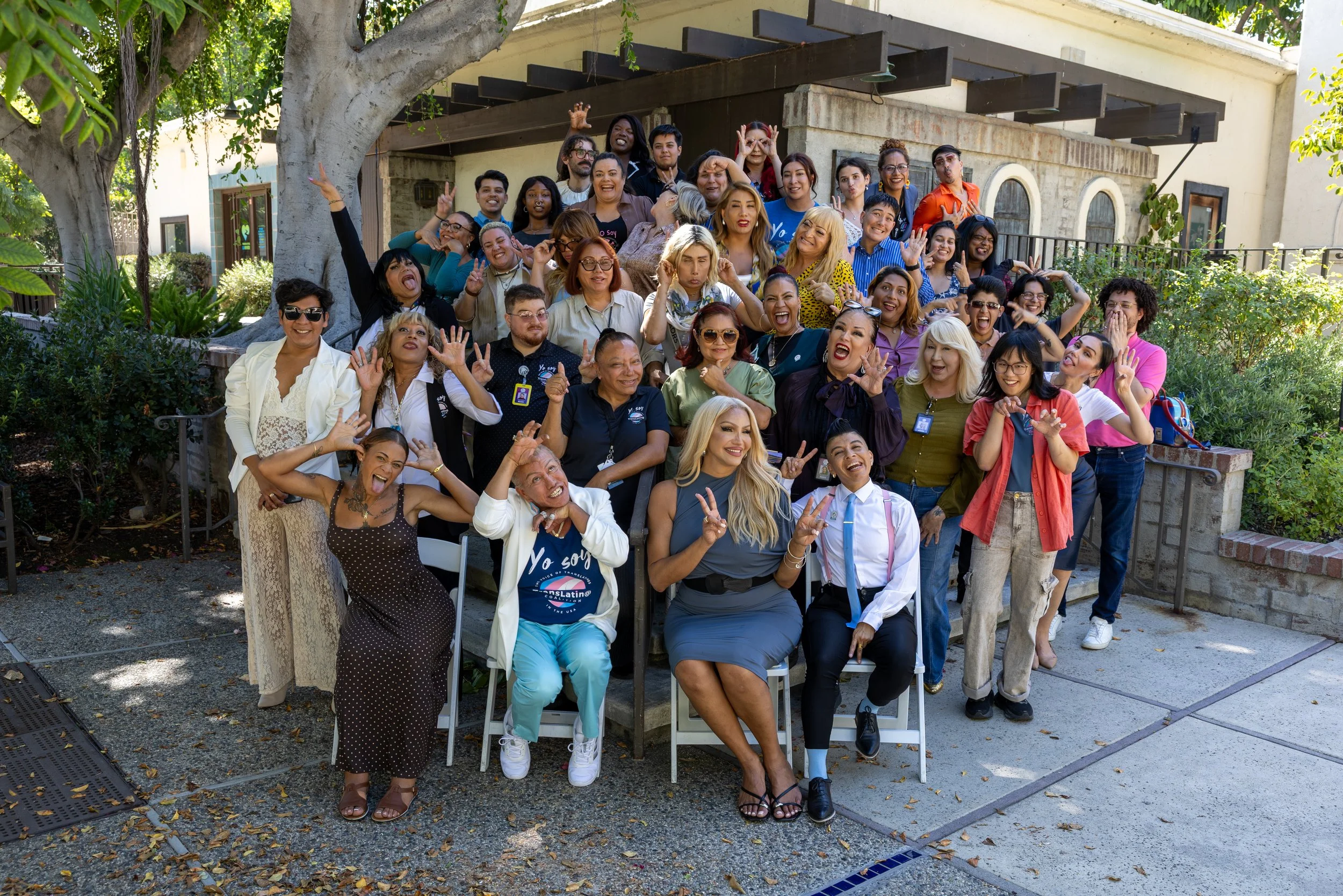 Un grupo de personas de todo tipo reunidas al aire libre frente a una casa rodeada de árboles, sonriendo, haciendo gestos divertidos y posando para una foto.