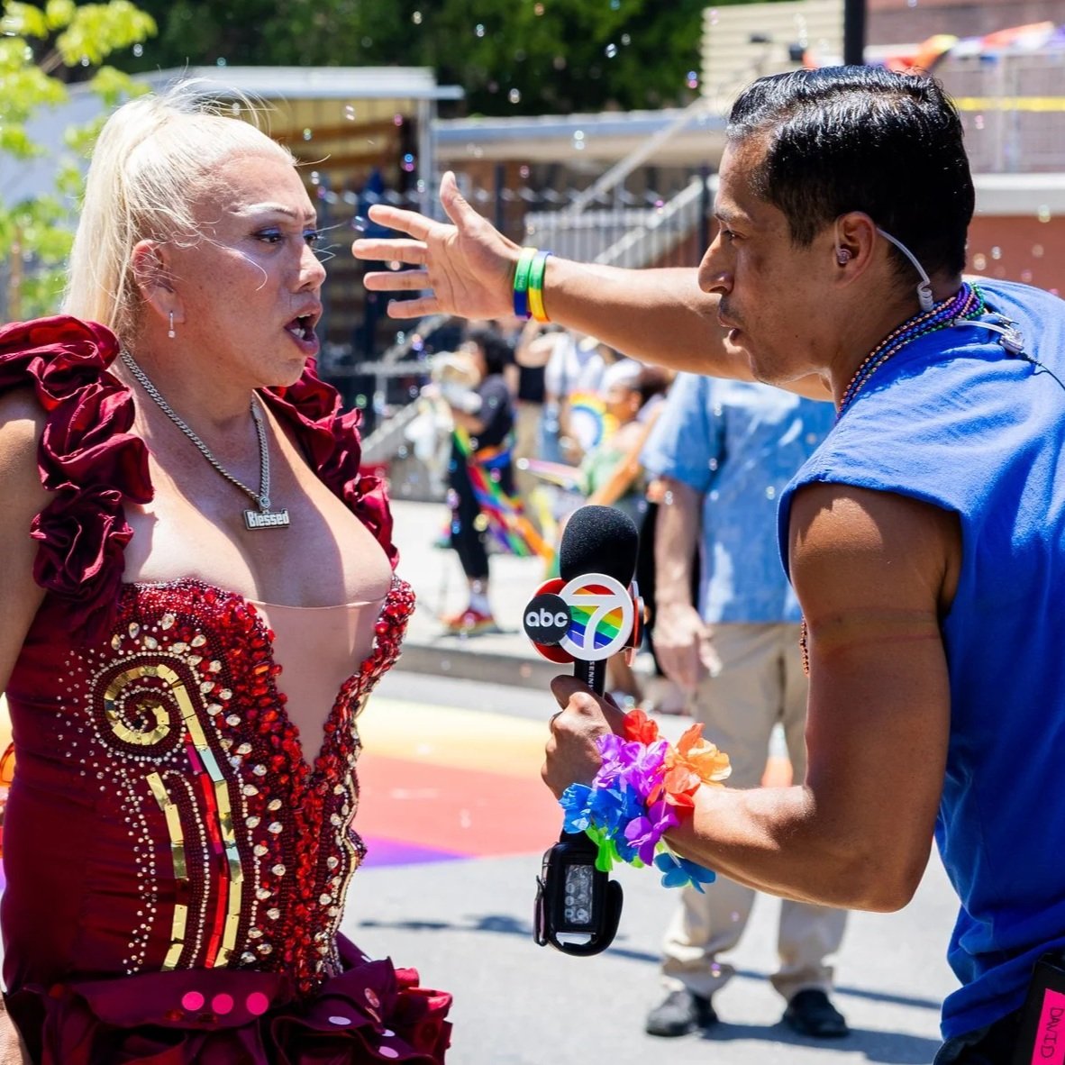 A woman in a red dress being interviewed by a male reporter at a pride event, with rainbow decorations and people in the background.