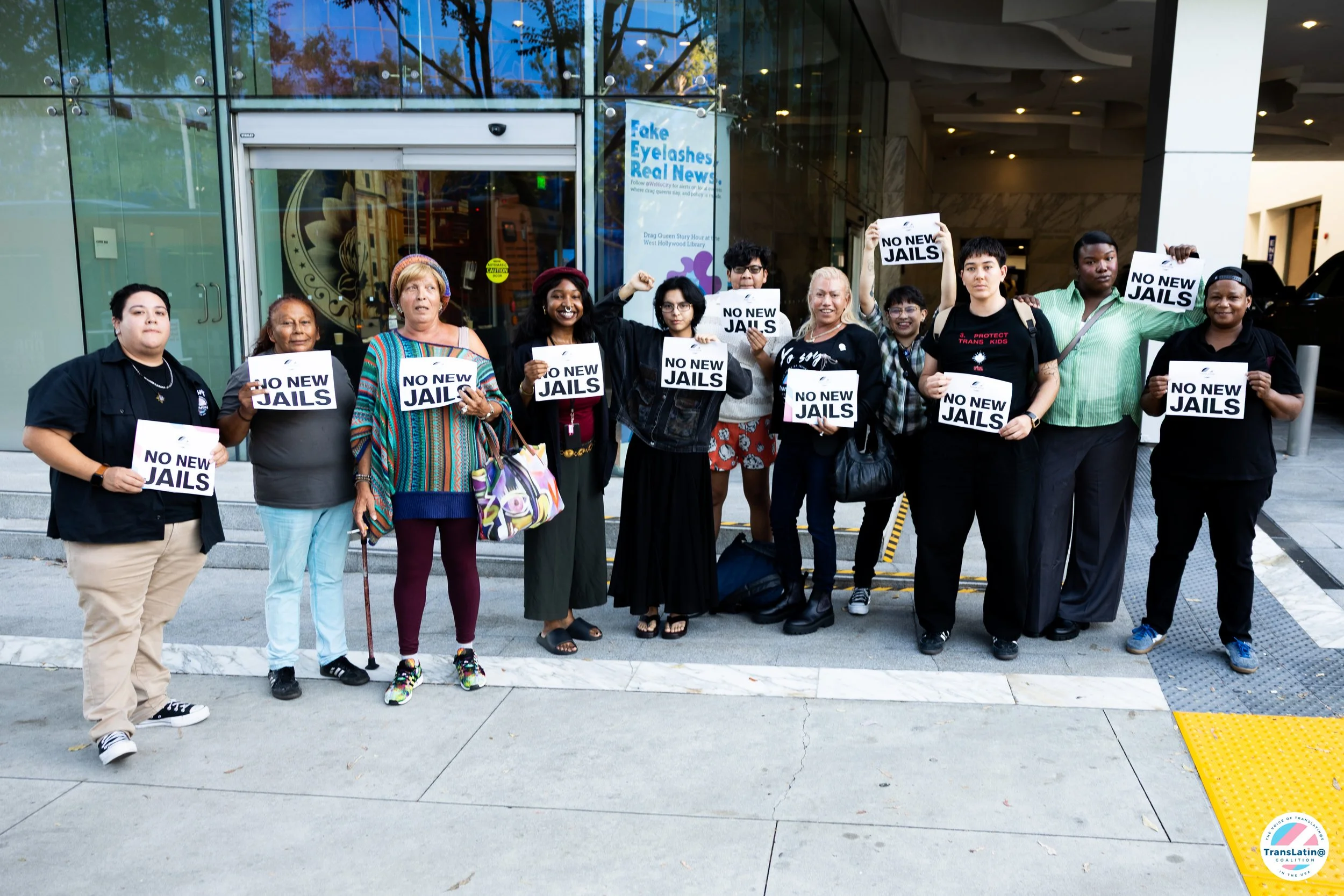 A group of community members stands outside the venue holding signs that read “No New Jails.”