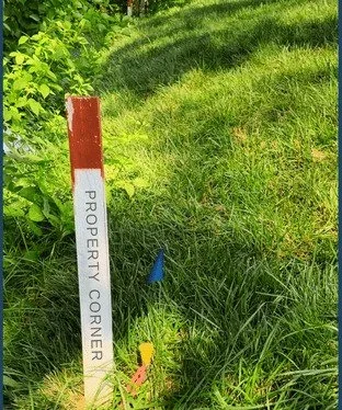 A white property corner survey stake marked “PROPERTY CORNER” standing in a grassy yard with survey flags, showing a property boundary relevant to fence planning by K&C Fence.