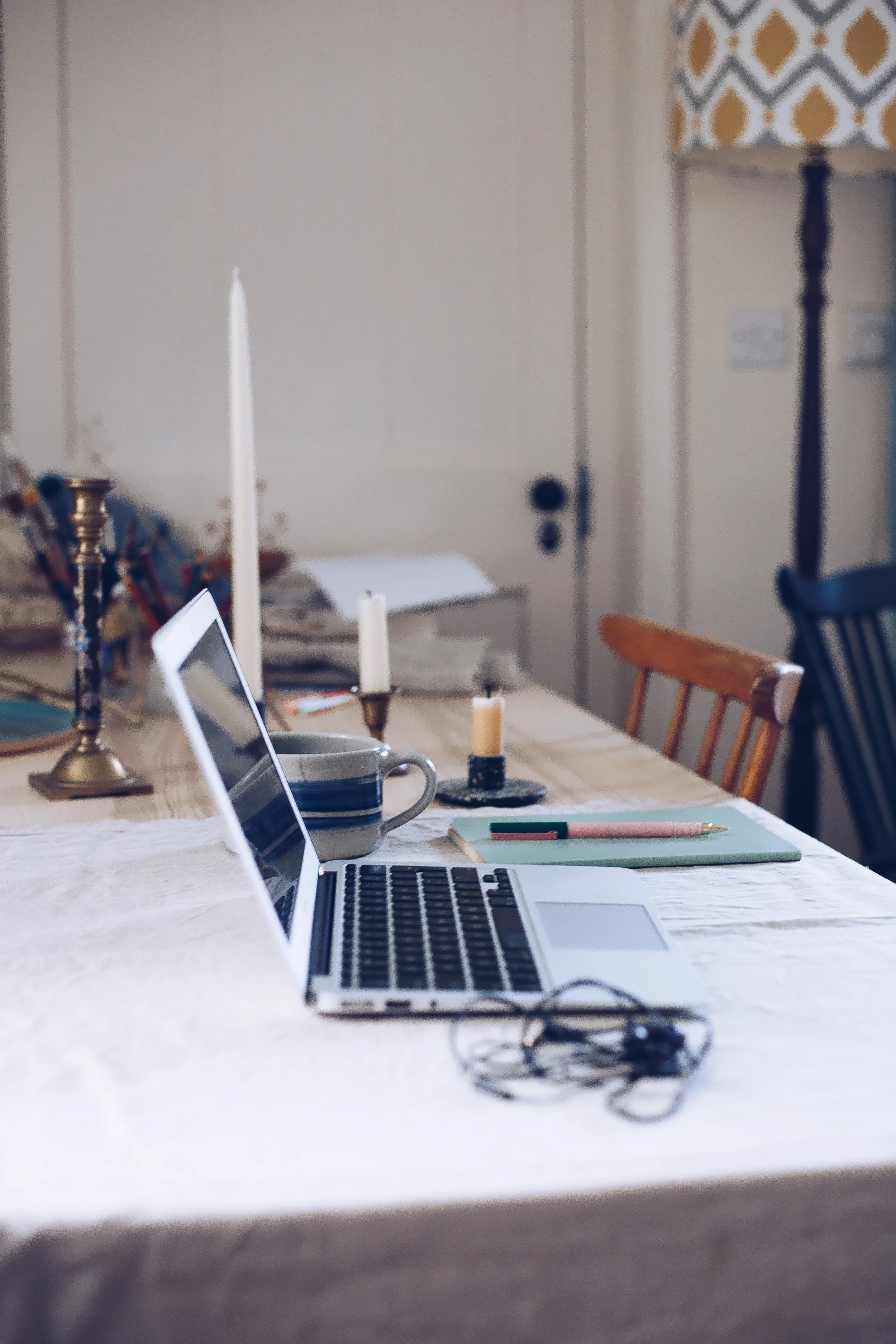 A dining table with a laptop, a cup, a notebook, a pen, and candles, set in a cozy room.