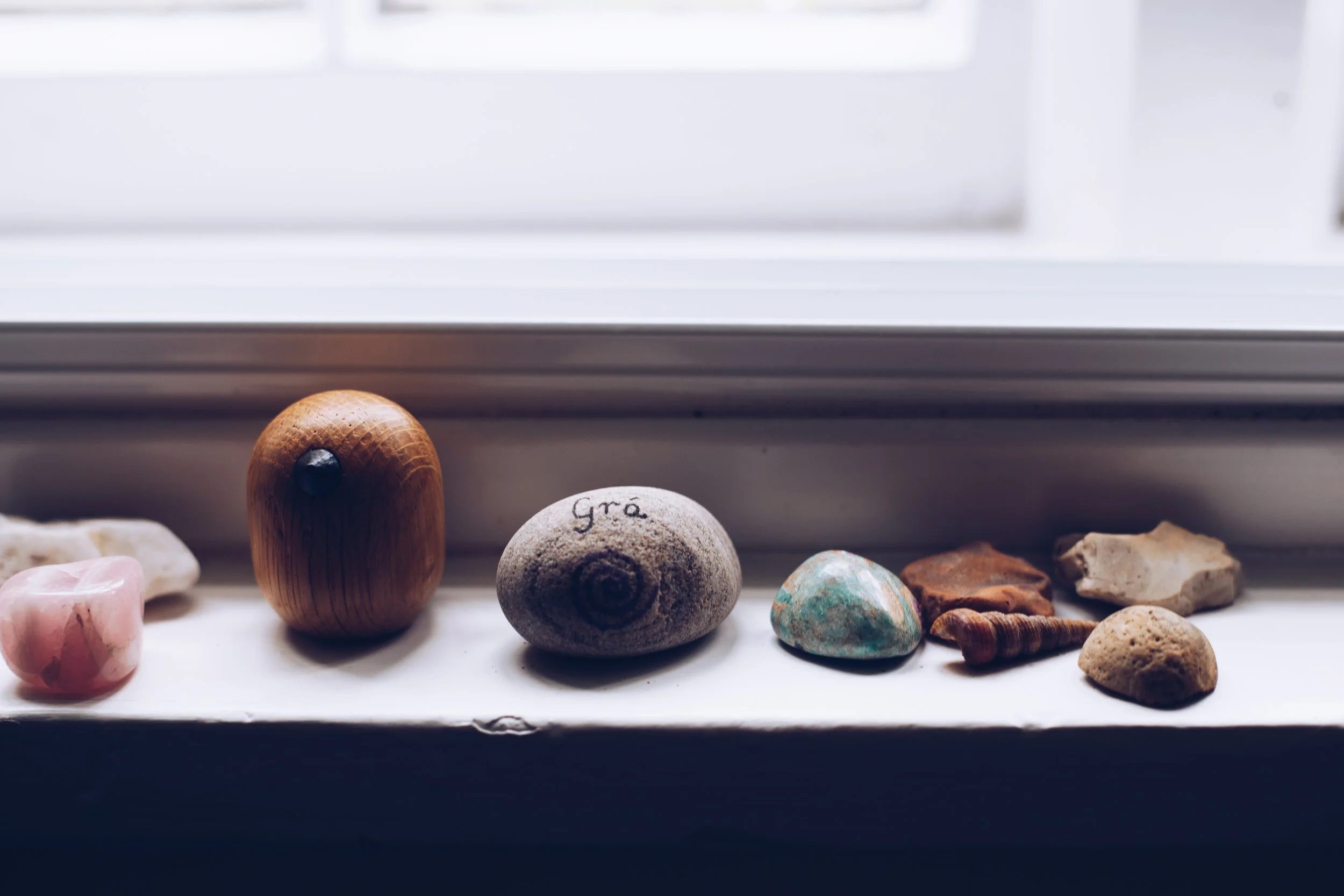A collection of small objects on a windowsill, including a pink stone, a wooden bead, a gray rock with writing, a turquoise stone, a layered rock, a brown shell, and a tan stone. The background shows a window with white framing.