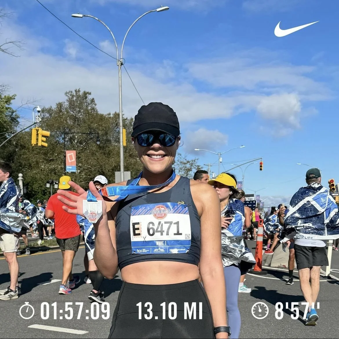 Female marathon runner holding medal at finish line, surrounded by other runners, with clear blue sky and streetlights in background, wearing sunglasses, black cap, athletic outfit, race bib number 6471, and showing hydration medal.