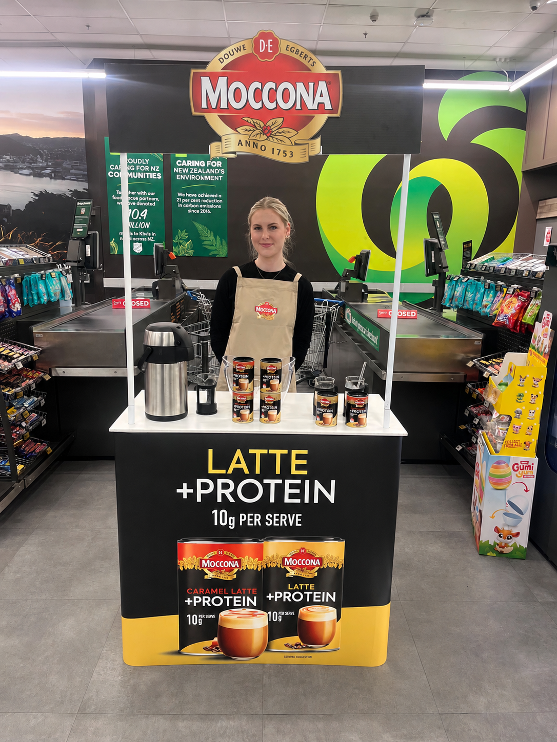 Promostaff grocery demonstrator.  Moccona coffee promotional stand with Latte + Protein drink cans, in a supermarket aisle.
