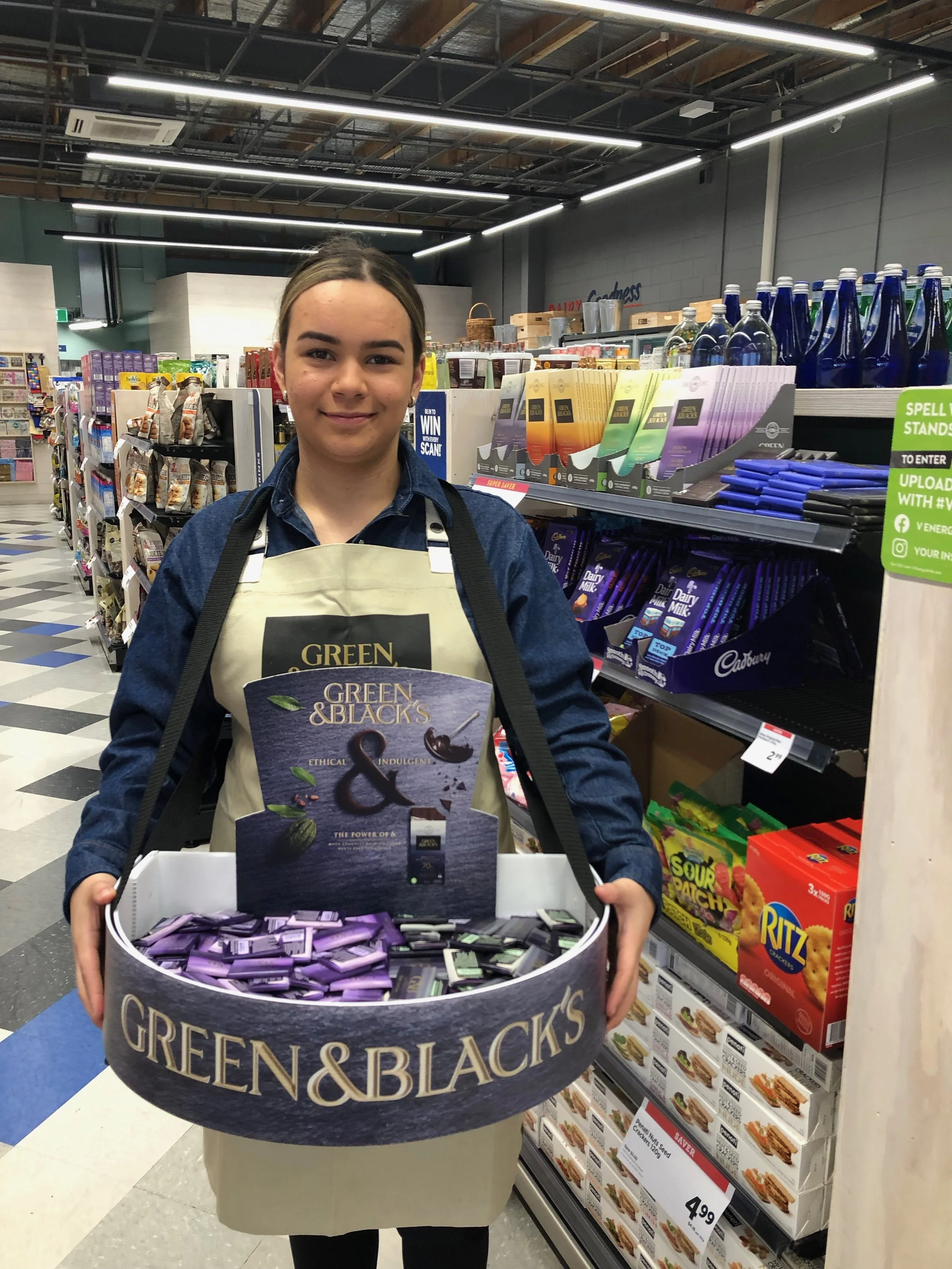 Promostaff in a grocery store, holding a large basket filled with Green & Black's chocolate bars.