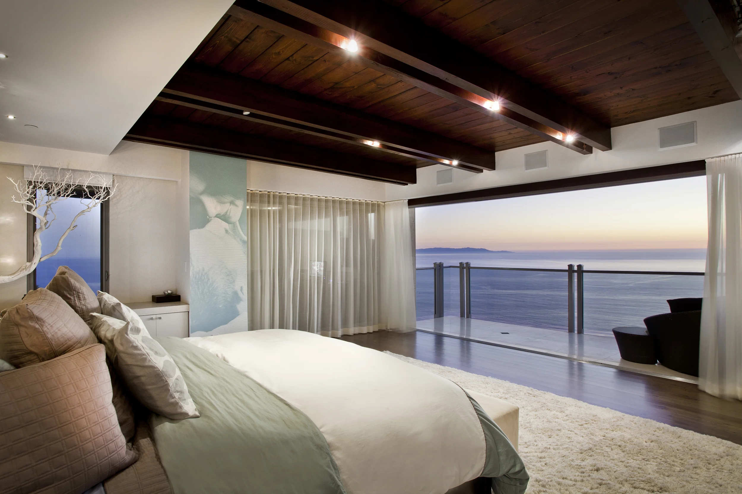A bedroom with a large bed, multiple pillows, a view of the ocean through sliding glass doors, and a balcony overlooking the water at sunset.