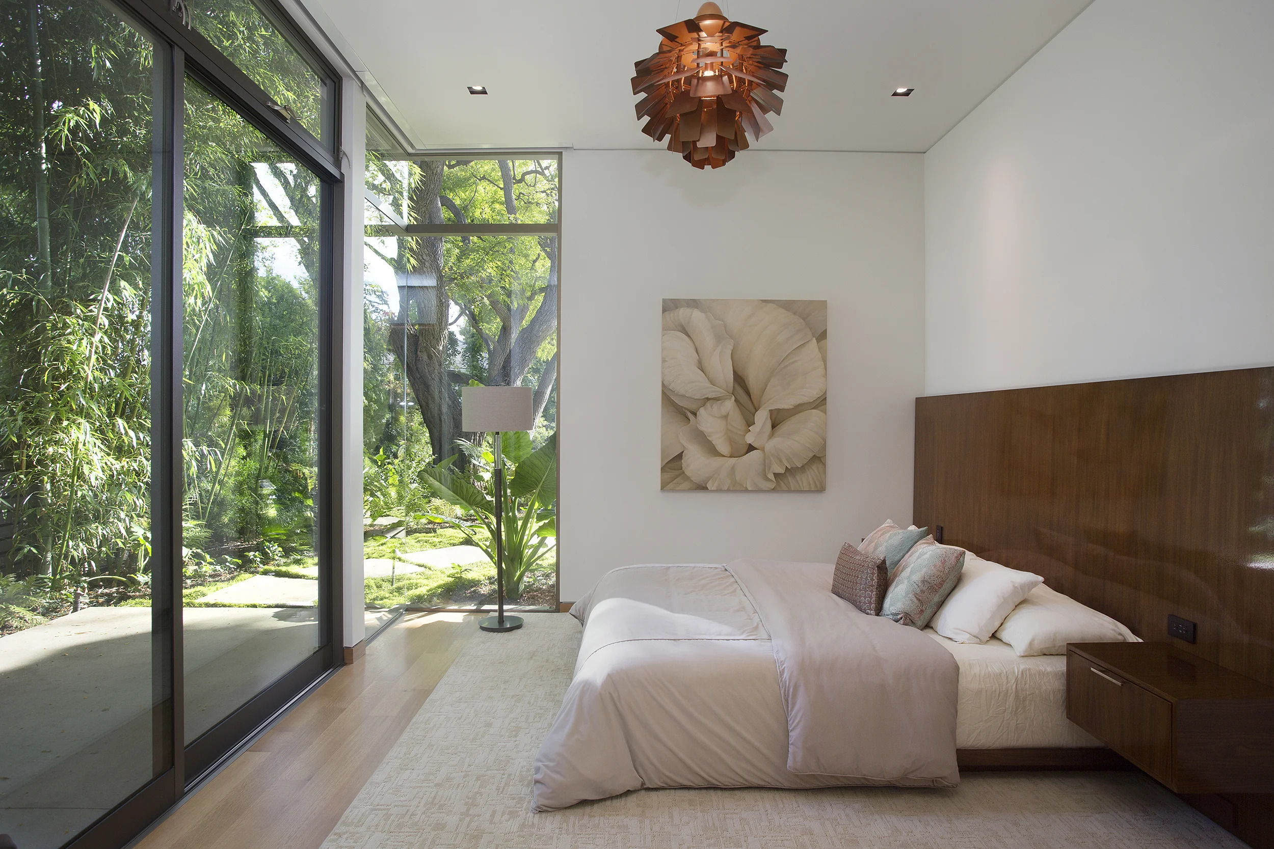 A modern bedroom with a large glass sliding door, a bed with a wooden headboard, a beige rug, a floor lamp, wall art, and a view of greenery outside.