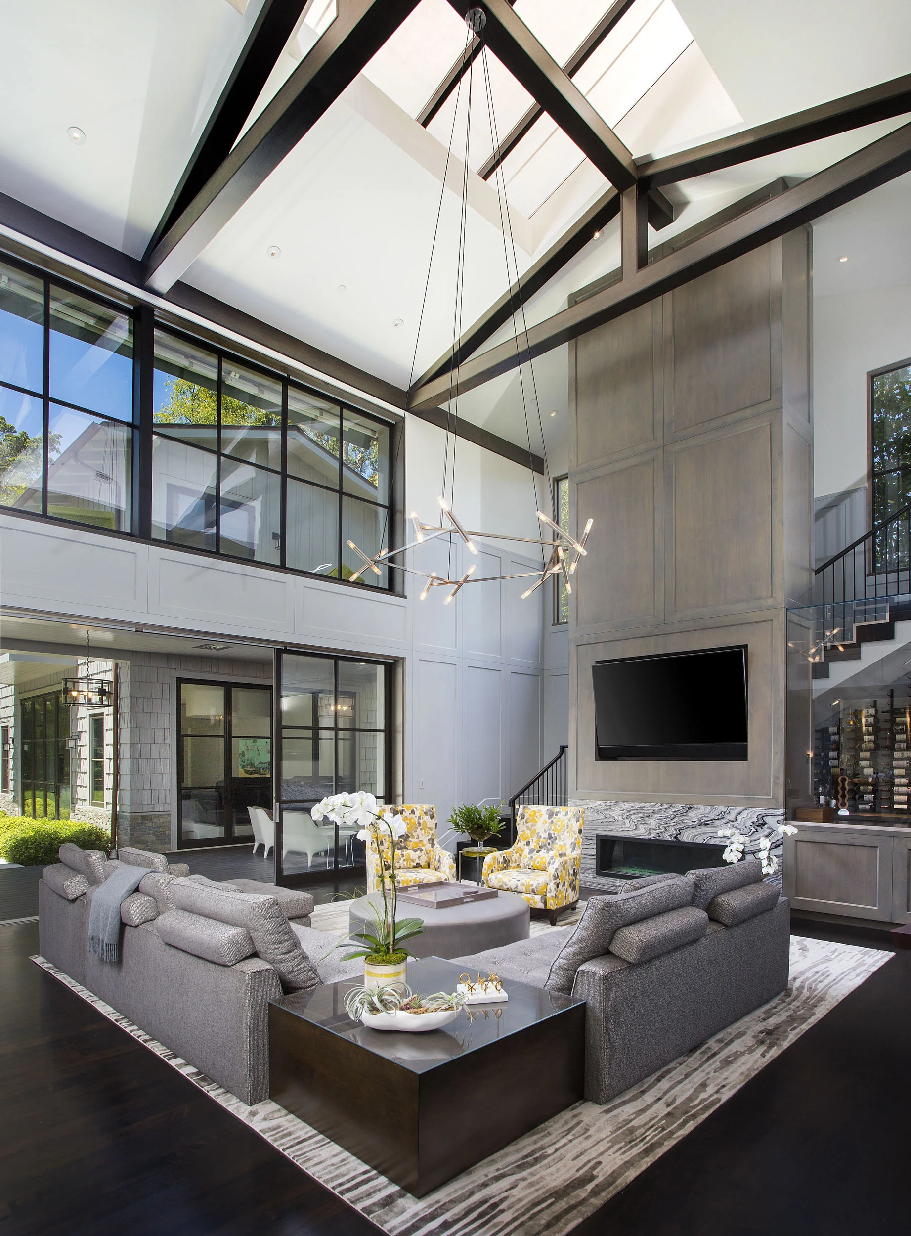 Modern living room with high ceiling, large glass windows, and a contemporary chandelier. There is a gray sofa with decorative pillows, two yellow floral armchairs, a round ottoman, and a black side table with a plant. The room has a fireplace with a
