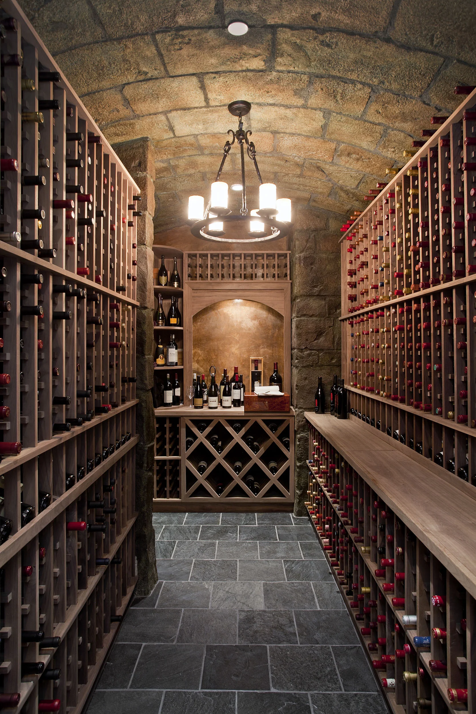 A wine cellar with stone walls and floor, wooden wine racks on both sides filled with bottles, and a wooden counter at the back displaying several bottles of wine. A chandelier with multiple bulbs hangs from the ceiling.