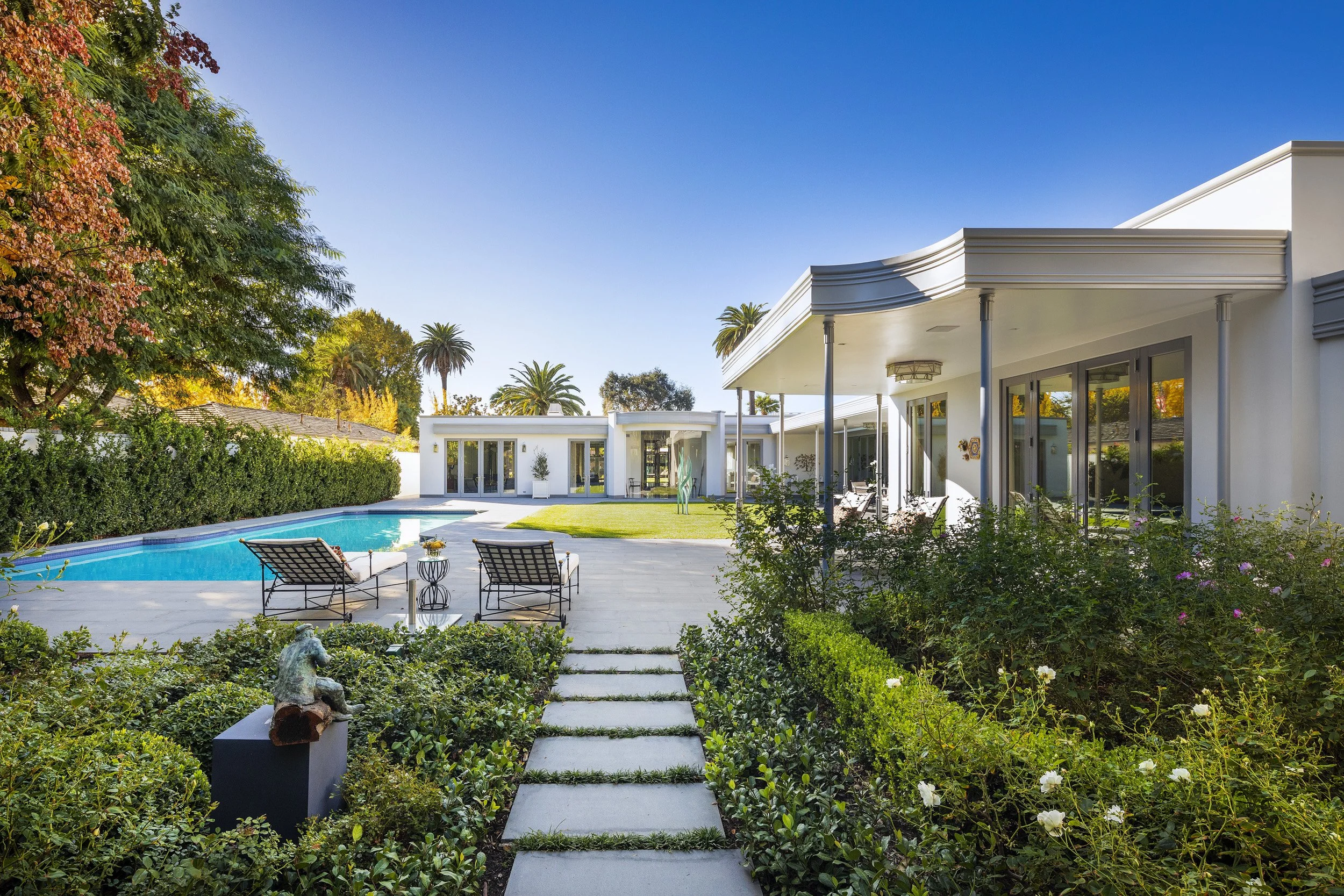 Modern white house with a backyard pool, surrounded by lush greenery and tall palm trees under a clear blue sky.