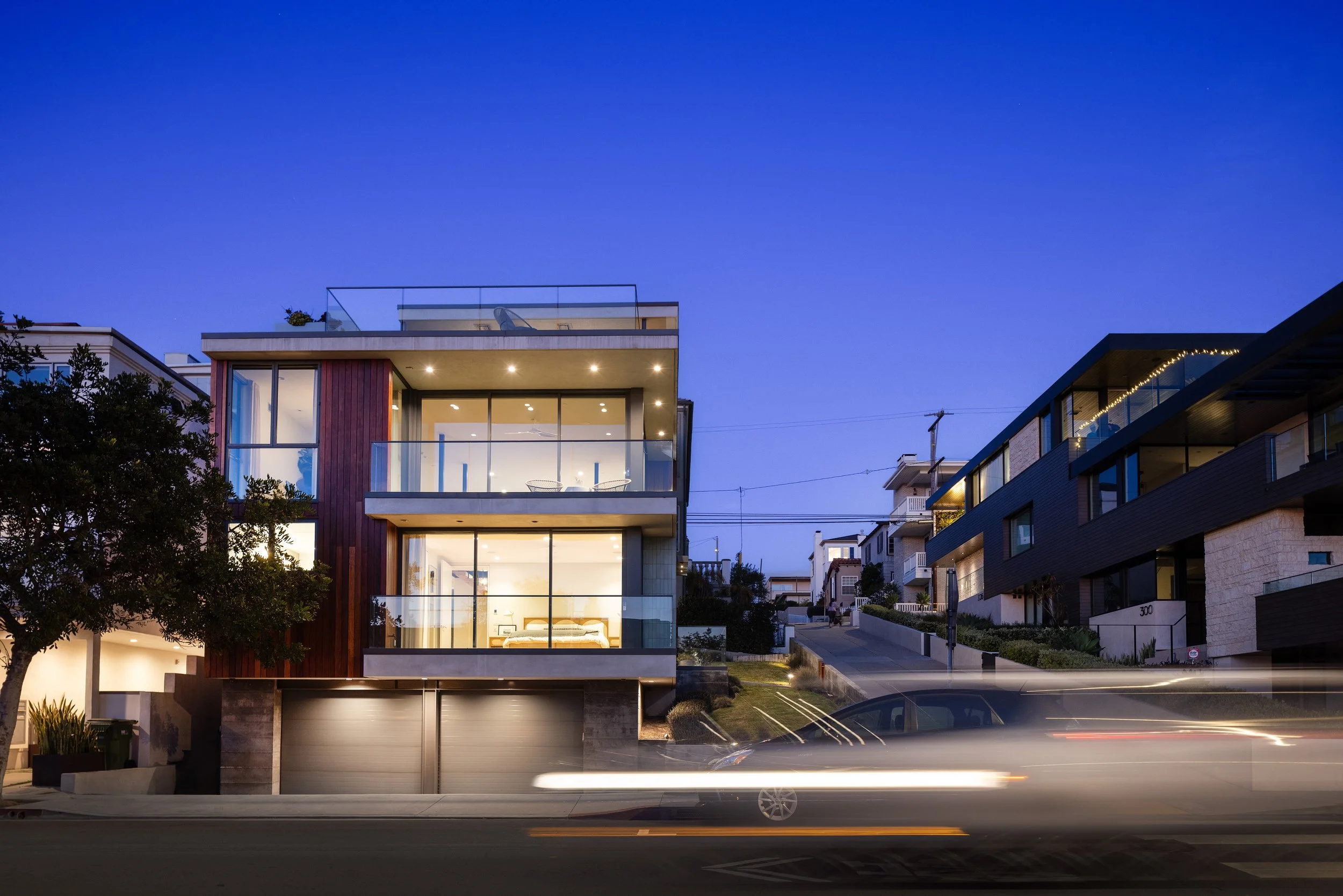 Modern multi-story houses on a hillside during twilight with glowing interior lights and a blurred moving car in the foreground.