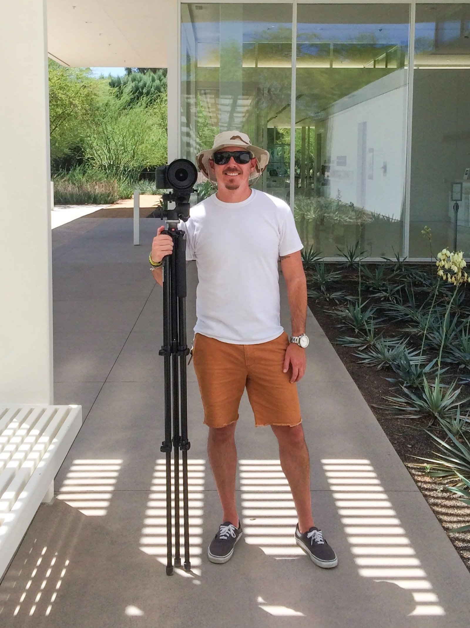 A man standing outdoors holding a camera on a tripod, wearing a wide-brimmed hat, sunglasses, a white t-shirt, brown shorts, and sneakers, with a modern building and greenery in the background.