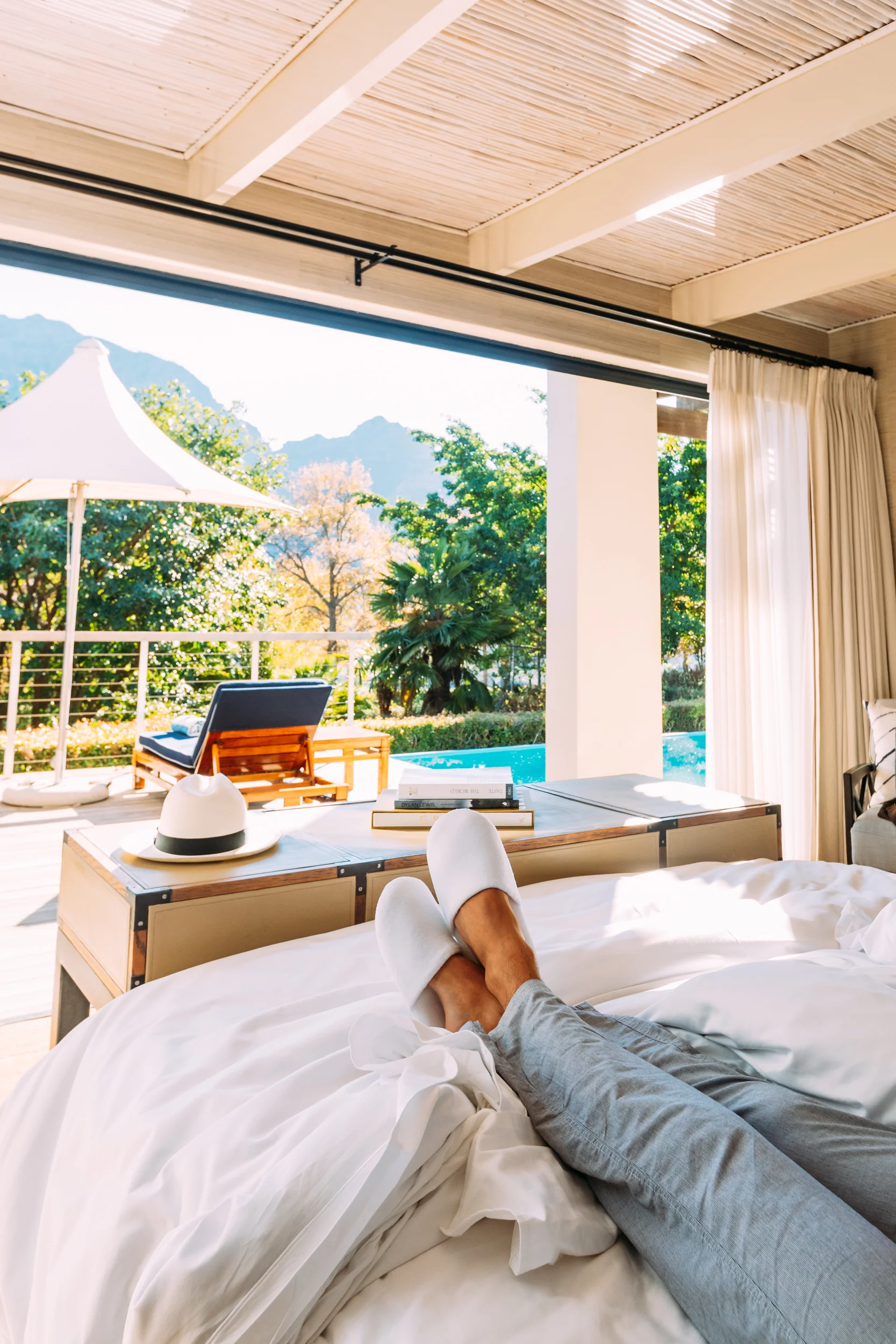A man with his feet up in bed with plush slippers enjoying the view from the Presidential Lodge at Delaire Graff Lodges & Spa