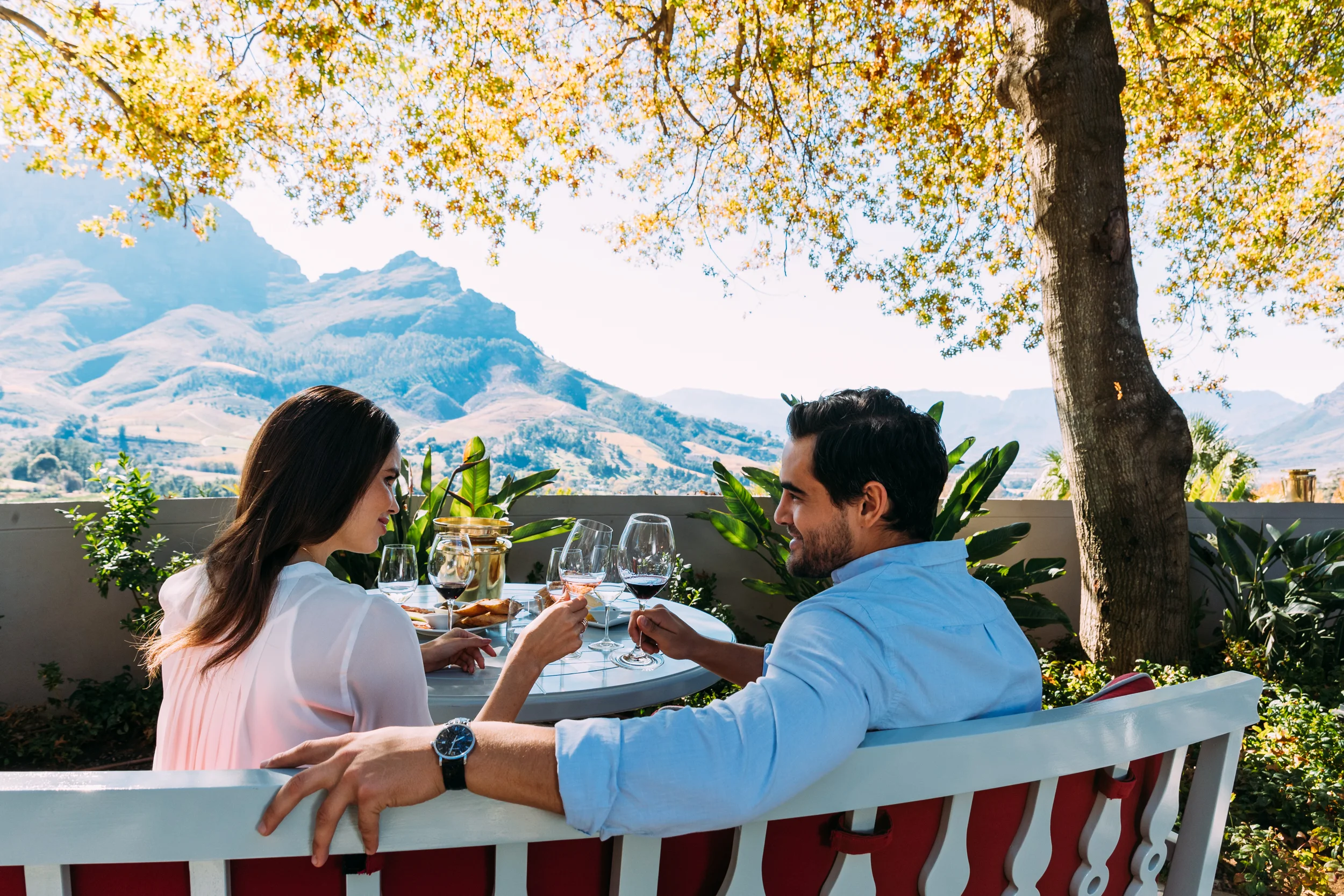 A couple clink glasses at the Wine Lounge at Delaire Graff Estate sitting at a table with a view of the mountains