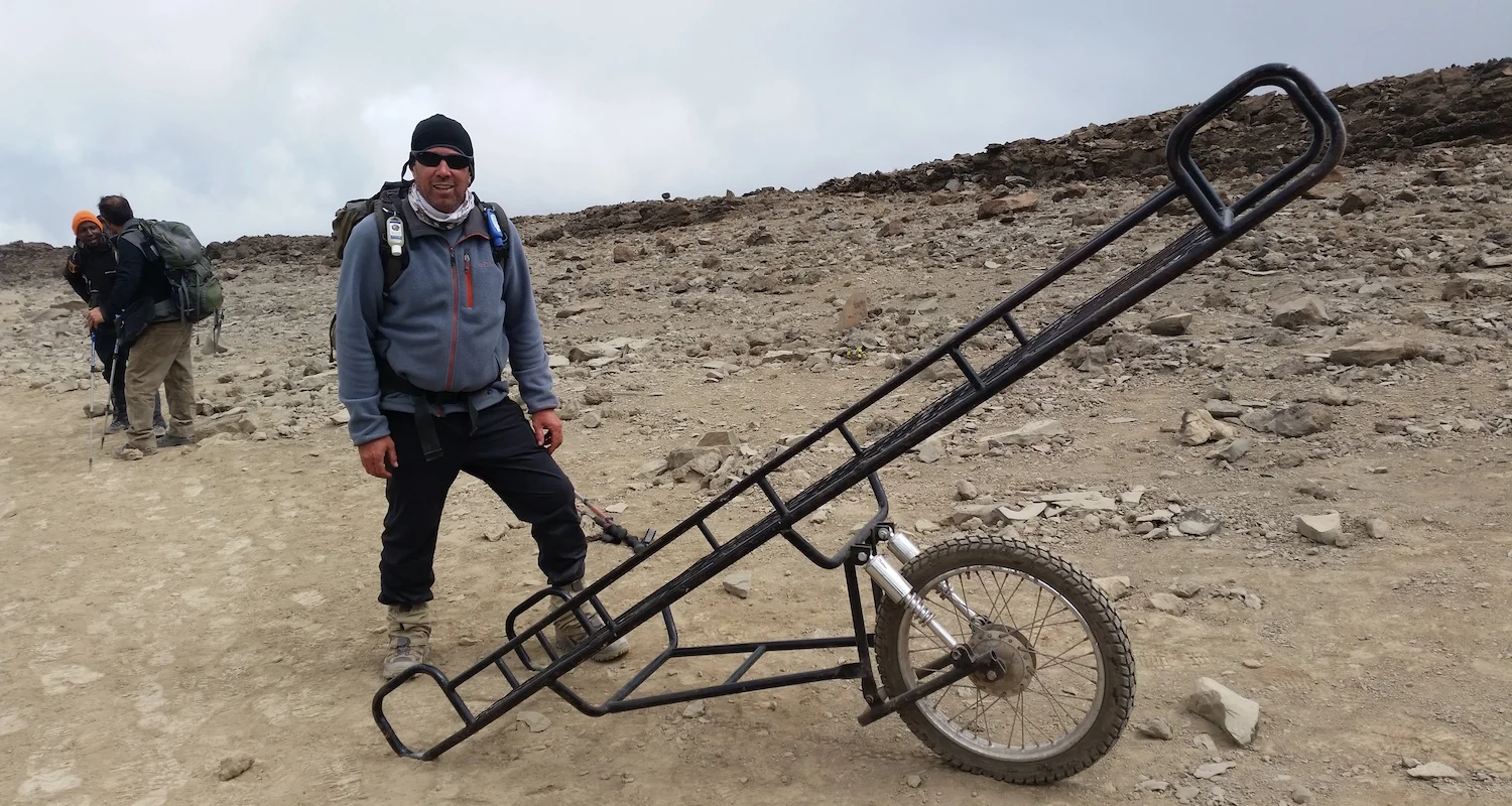 Tommy stands beside a "Kili cart" This unicycle "vehicle" is used to carry/roll injured climbers over rugged terrain.