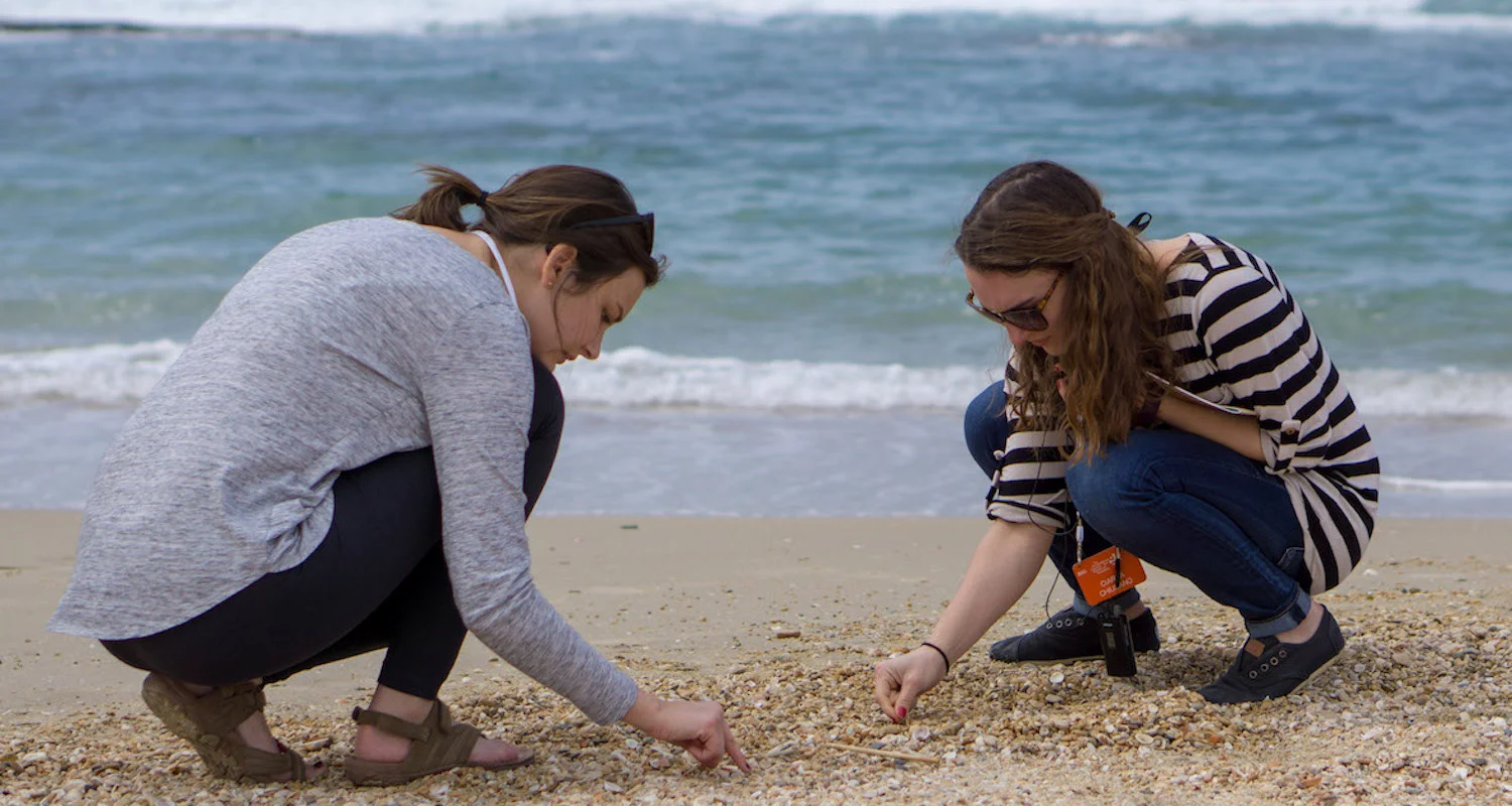 Michaela Wood (L) and Ciarra Chiusano (R) sort through seashells at Caesarea Maritima. The breakers of the Mediterranean Sea roll in softly behind them. Photograph by Bible Land Explorer Adrienne Griffin.