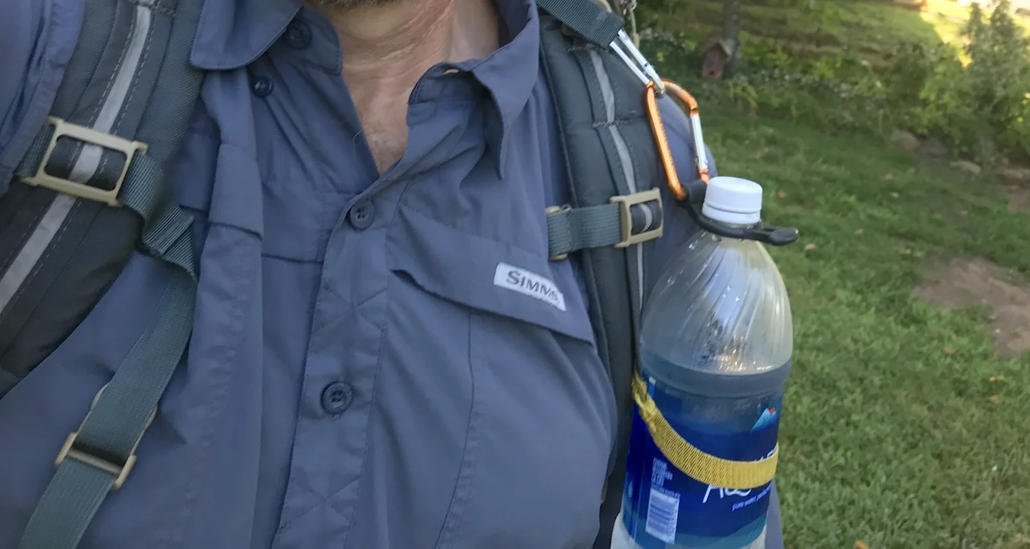 The Bottle Bandit makes it possible to clip a disposable water bottle to the shoulder strap of a backpack. The Bandit is made of black silicone. It fits tightly around the bottle neck just under the cap. The hack was the elastic band (yellow in color here) that held the bottle securely against my body.