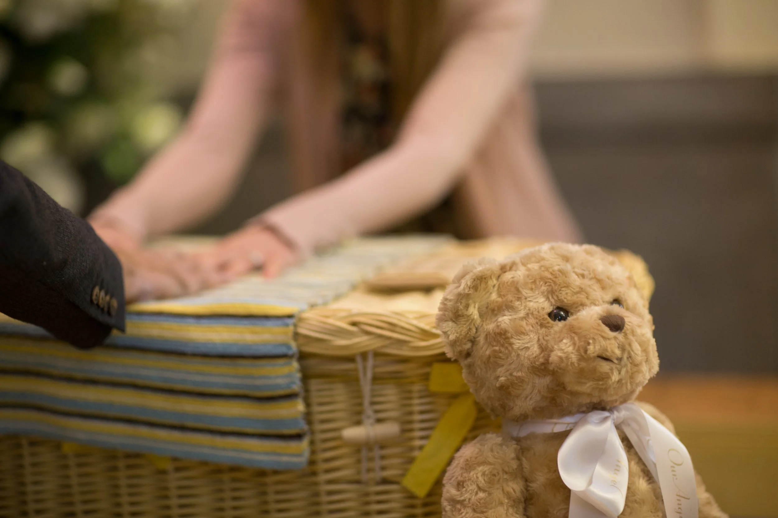 A teddy bear with a white ribbon beside a small woven coffin