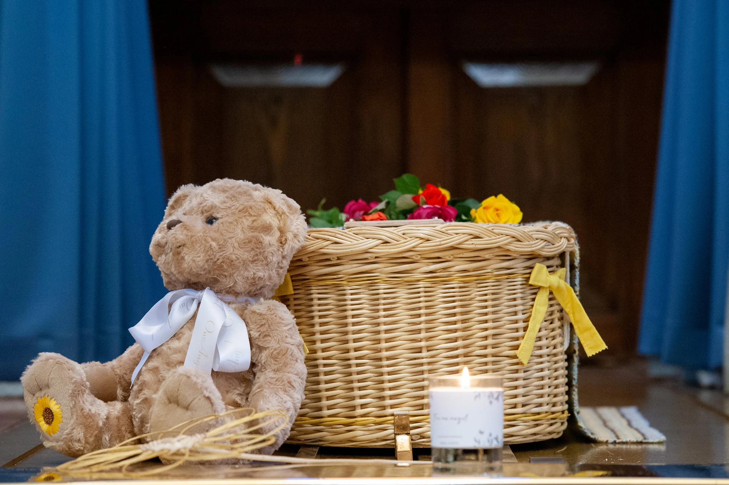 A teddy bear, candle and flowers beside a small woven coffin