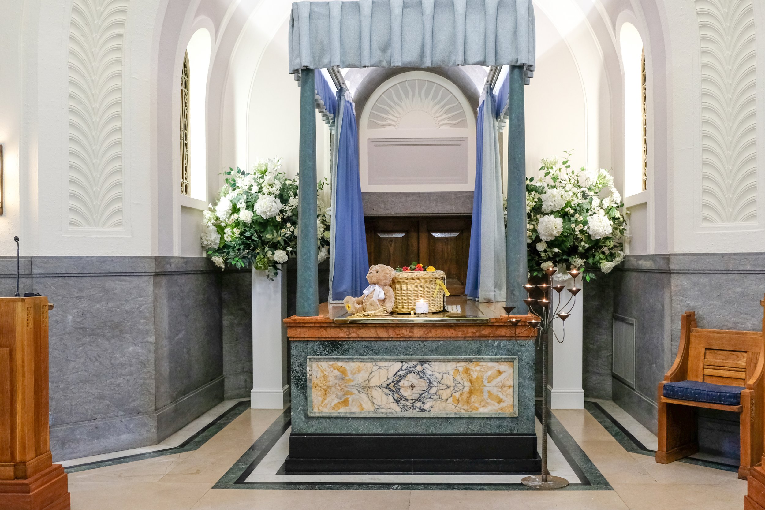 A small wicker coffin on a chapel catafalque with white flowers beside it